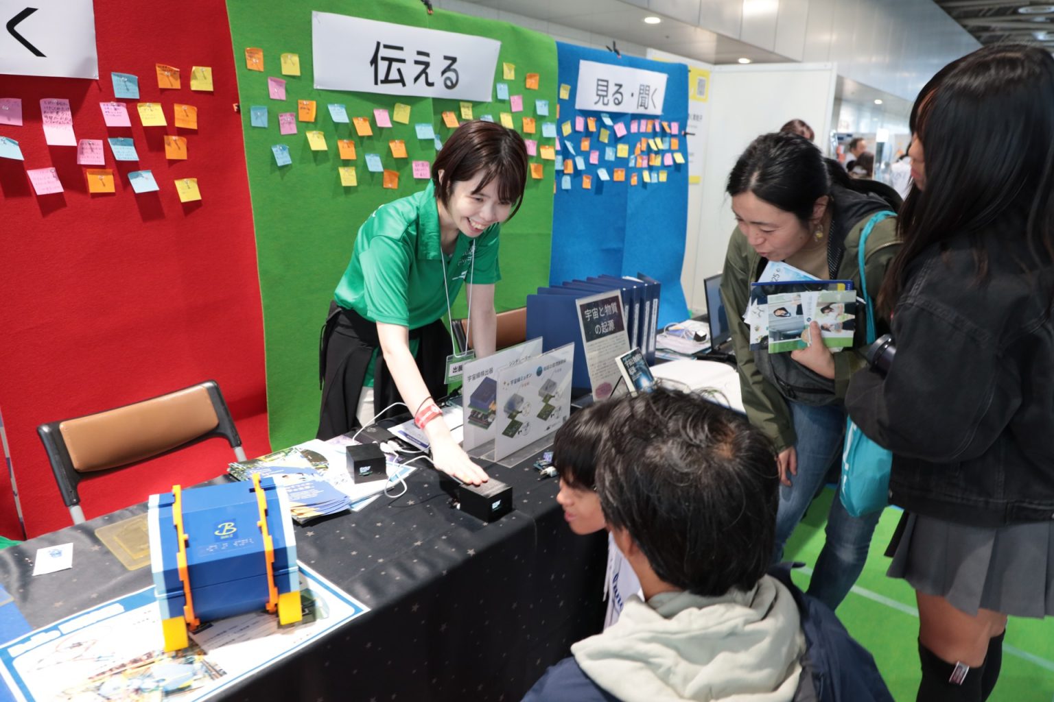 サイエンスアゴラ2025のKEK×加速キッチン共同ブースで、宇宙線検出器を手に来場者へ説明するスタッフ