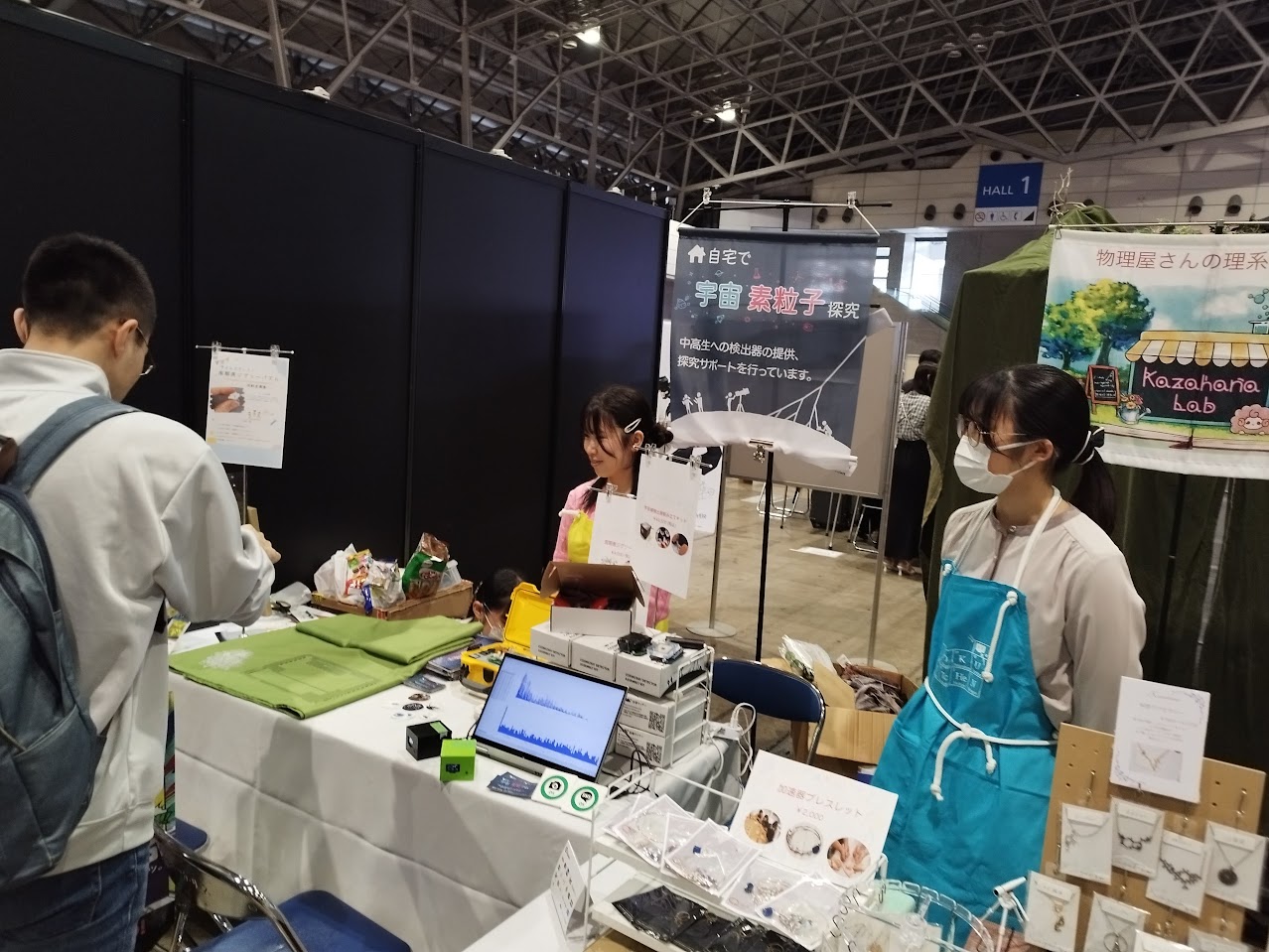 ニコニコ超会議2025の加速キッチンブースで、霧箱を並べたテーブル越しに来場者へ説明するスタッフ