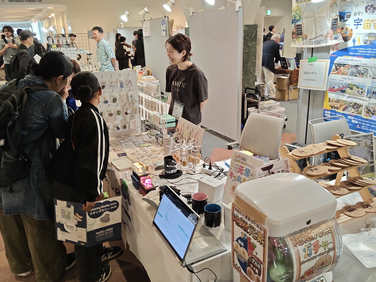 JpGU2026の加速キッチンブースで、宇宙線の測定画面や元素グッズを前に来場者と話すスタッフ