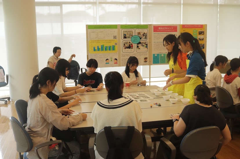 KEK一般公開2024で、ポスター展示を背にテーブルを囲んで実験に取り組む参加者