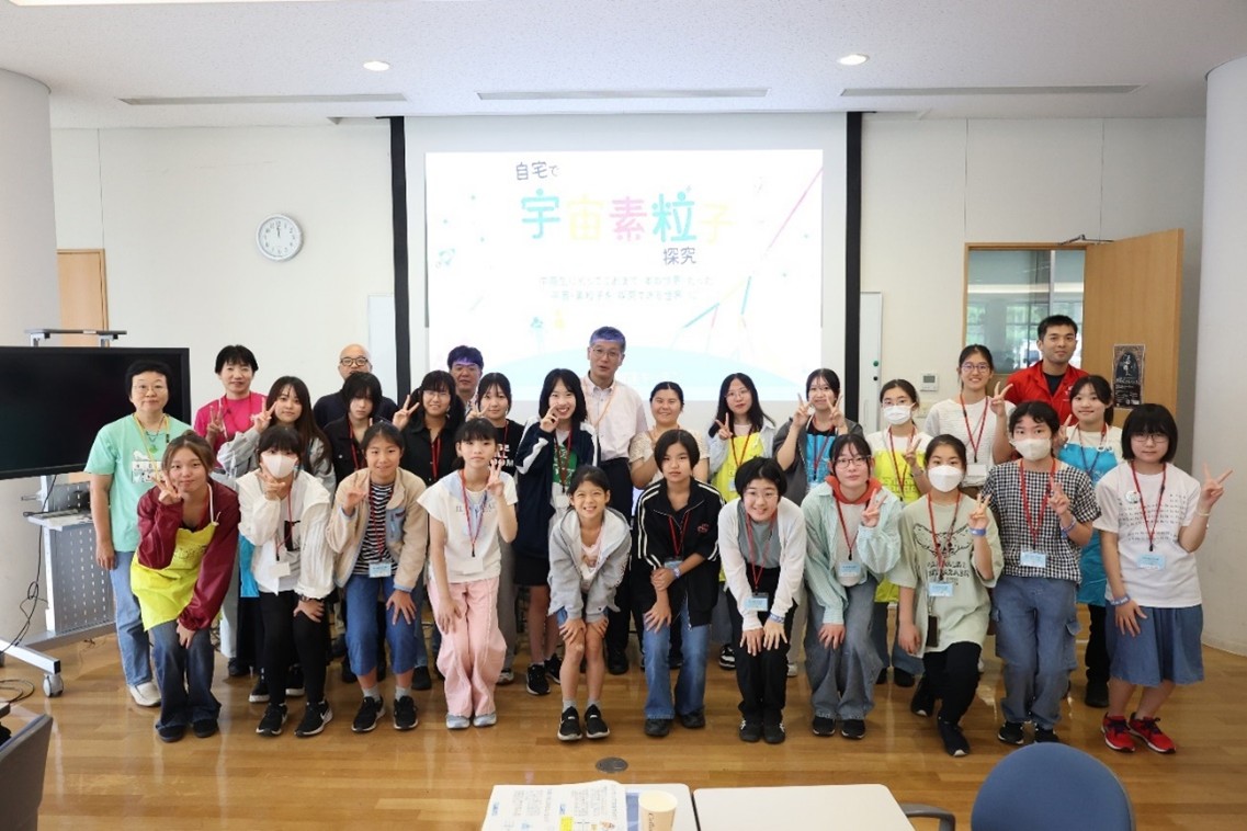 KEK一般公開2025の理系女子ミニツアー＆講演会で、「自宅で宇宙素粒子探究」のスライドを背に並ぶ参加者と研究者