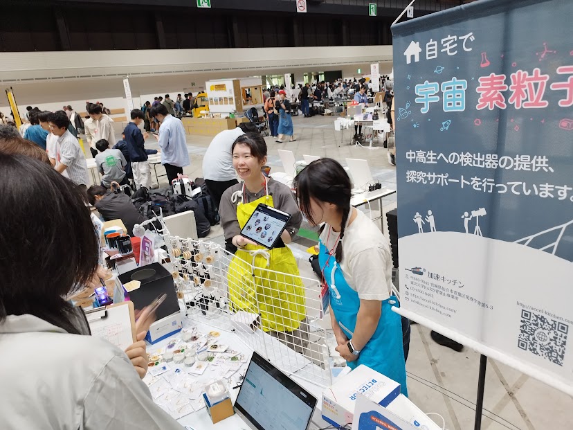 Maker Faire Tokyo 2026の加速キッチンブースで、タブレットや検出器を手に来場者へ説明するメンバーと「自宅で宇宙素粒子探究」のバナー