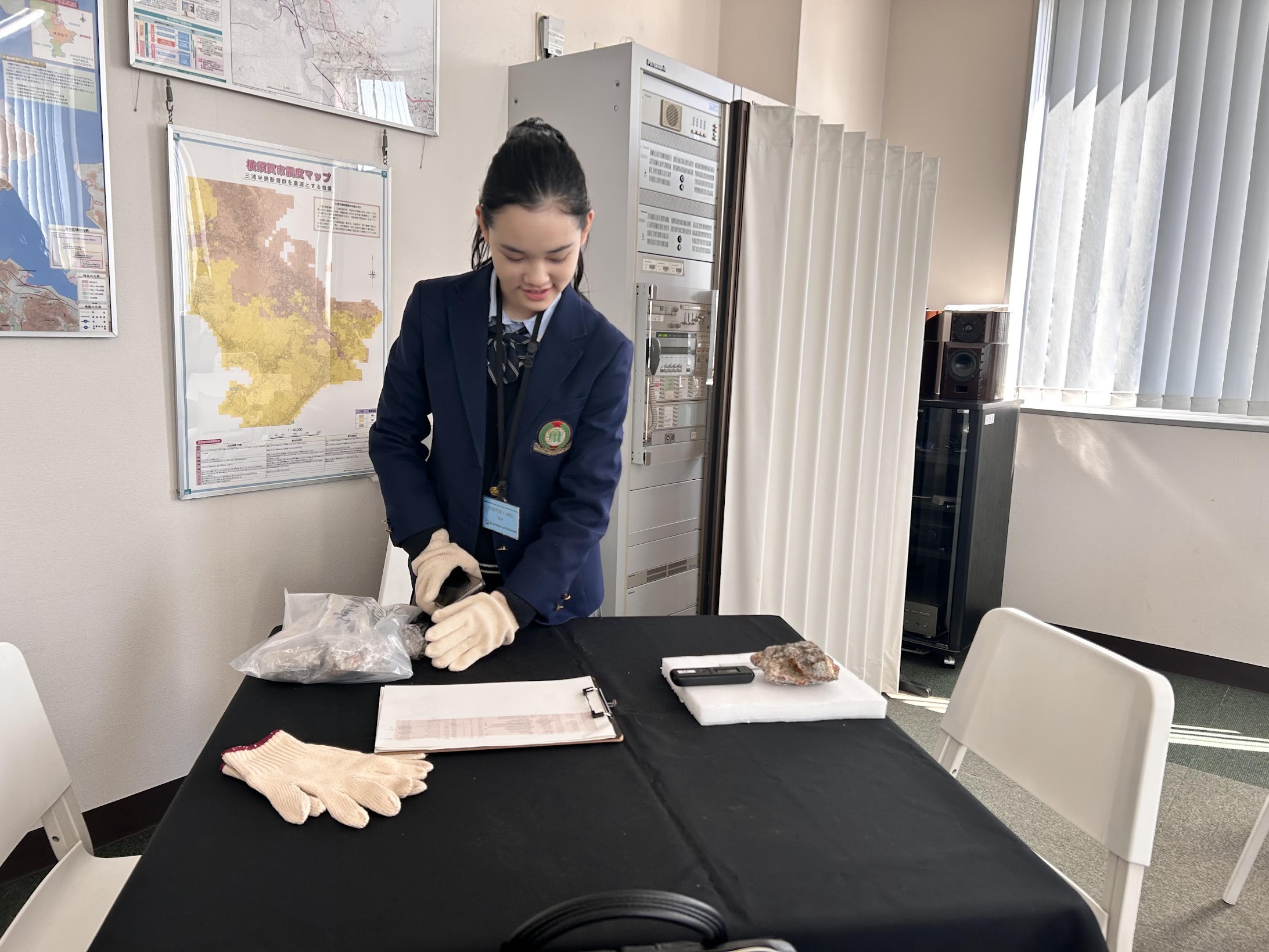 白い手袋をつけて岩石サンプルを扱う制服姿の生徒。背景に放射線測定器のラックと線量マップのポスター