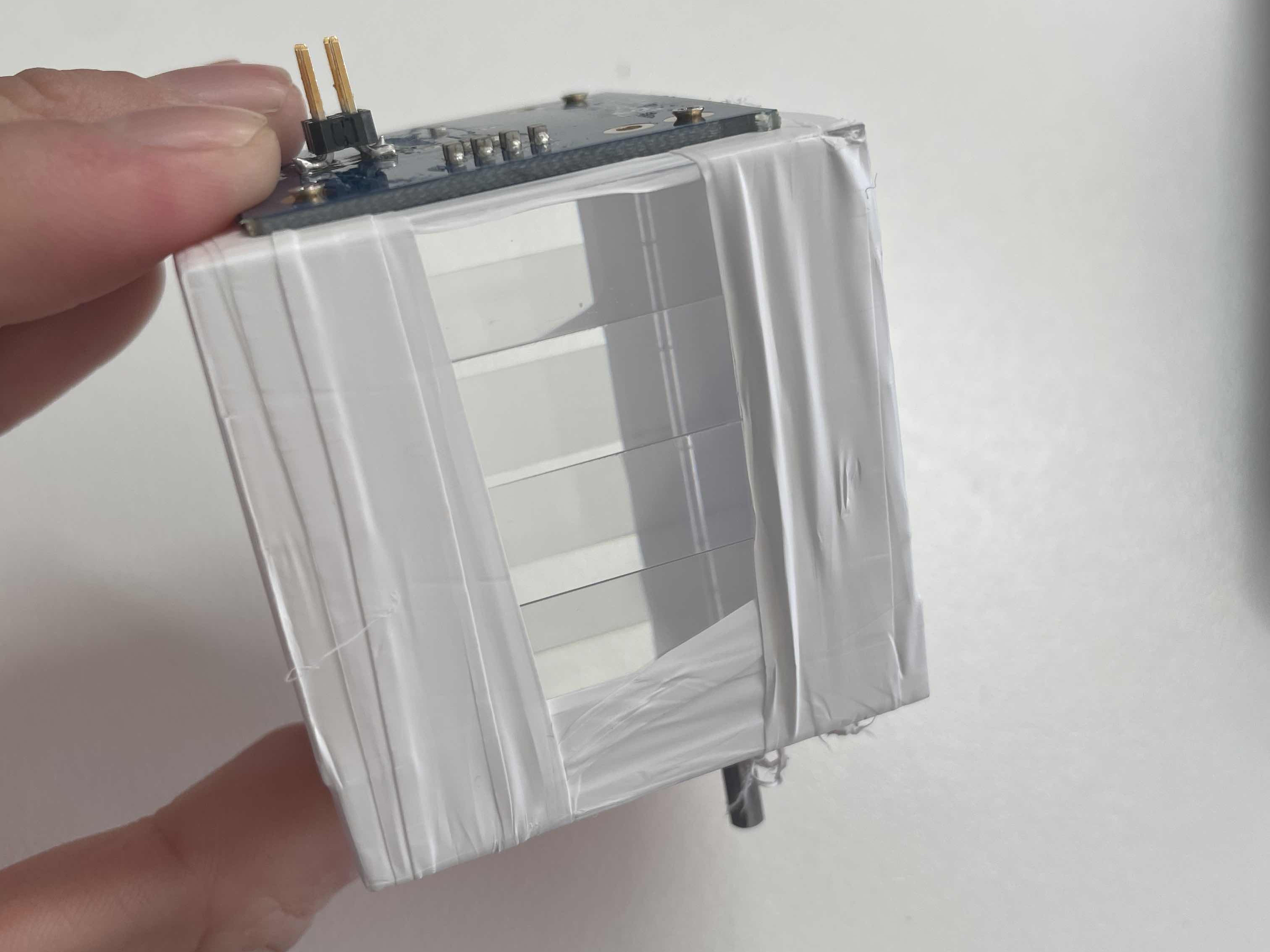 Hand holding a segmented detector: a stack of scintillator layers wrapped in white reflective tape, with the readout board on top