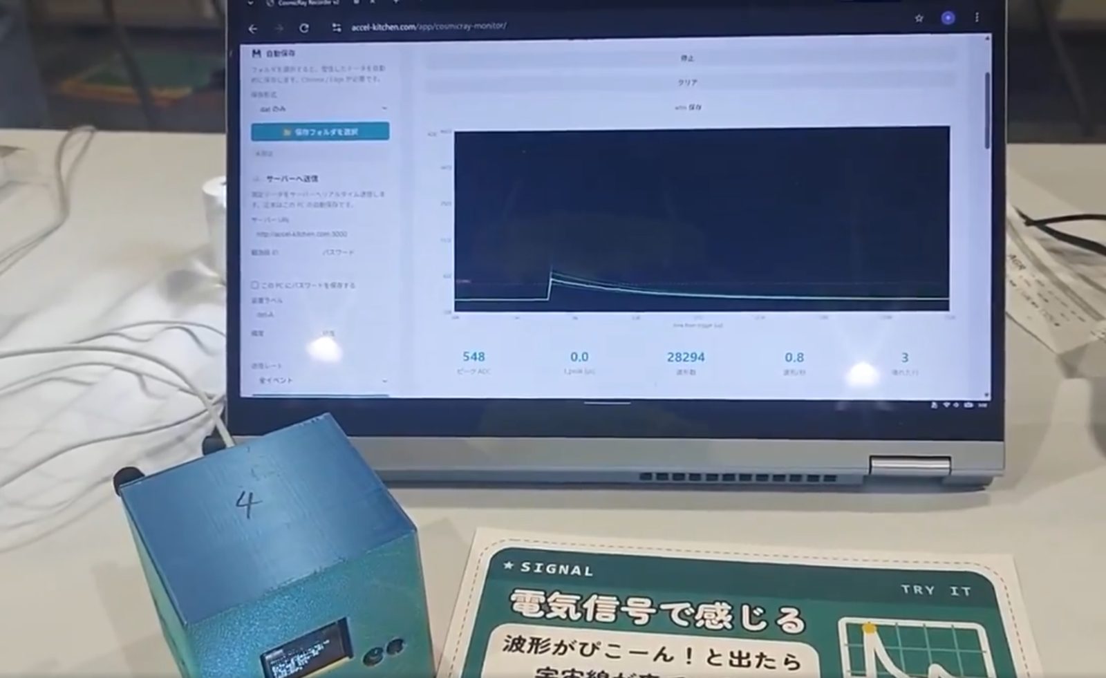 A laptop showing a cosmic-ray waveform and its peak ADC value, with a detector and a station card in front of it