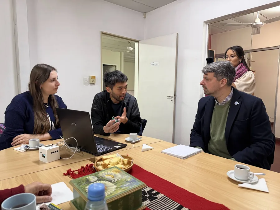 Five people seated around a meeting table. Kazuo Tanaka holds up a palm-sized detector as he explains it to a man in a jacket across the table; a laptop and a white detector box sit to his left.