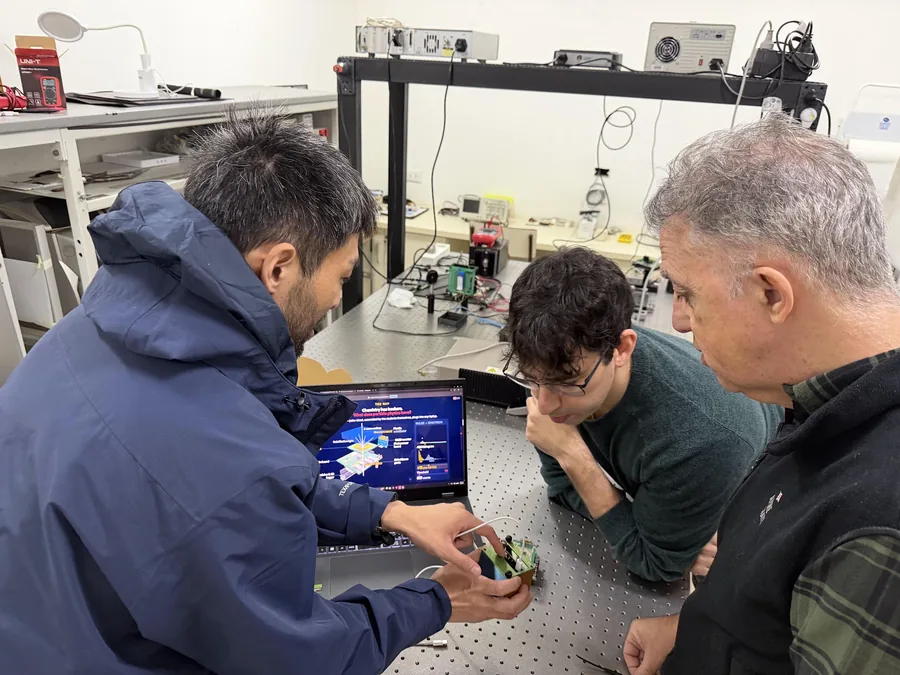 光学実験室の除振台の前で、田中香津生が基板がむき出しの検出器を手に持ち、二人の研究者が身を乗り出してのぞき込んでいる。奥にはレーザーの光学系が並ぶ