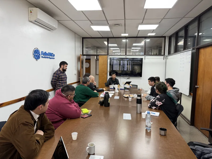 工学部の会議室。壁に FaIn·UNCo のロゴがあり、長机を囲んだ十数人の学生・教員に向けて、正面に座った田中香津生がスクリーンを使って話している