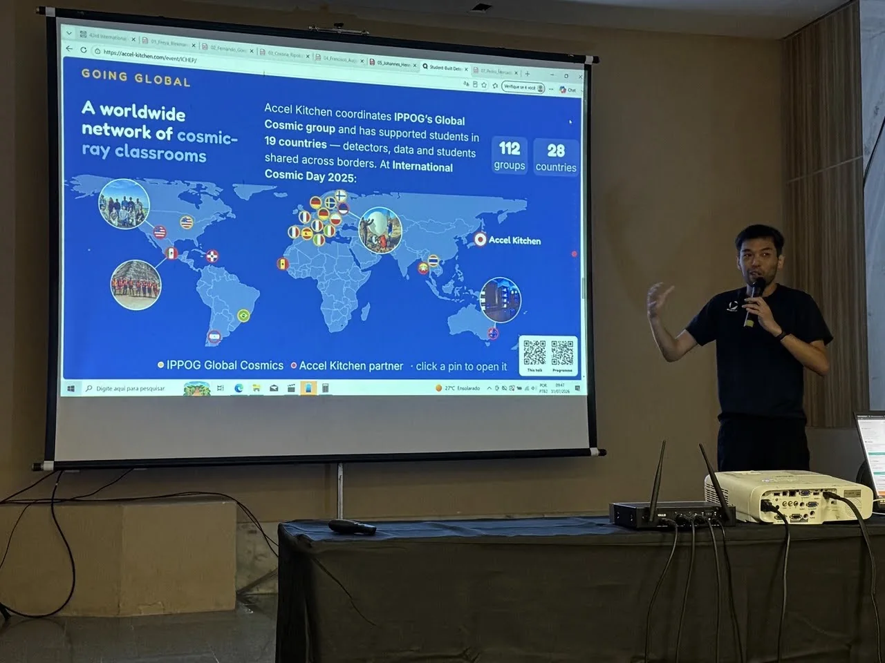 ICHEP2026 の会場で、スクリーンの横に立ちマイクを持って話す田中香津生。スクリーンには「A worldwide network of cosmic-ray classrooms」と題した青い世界地図のスライドが映り、各国の参加グループの写真と国旗が配置され、右上に「112 groups」「28 countries」の数字が並んでいる