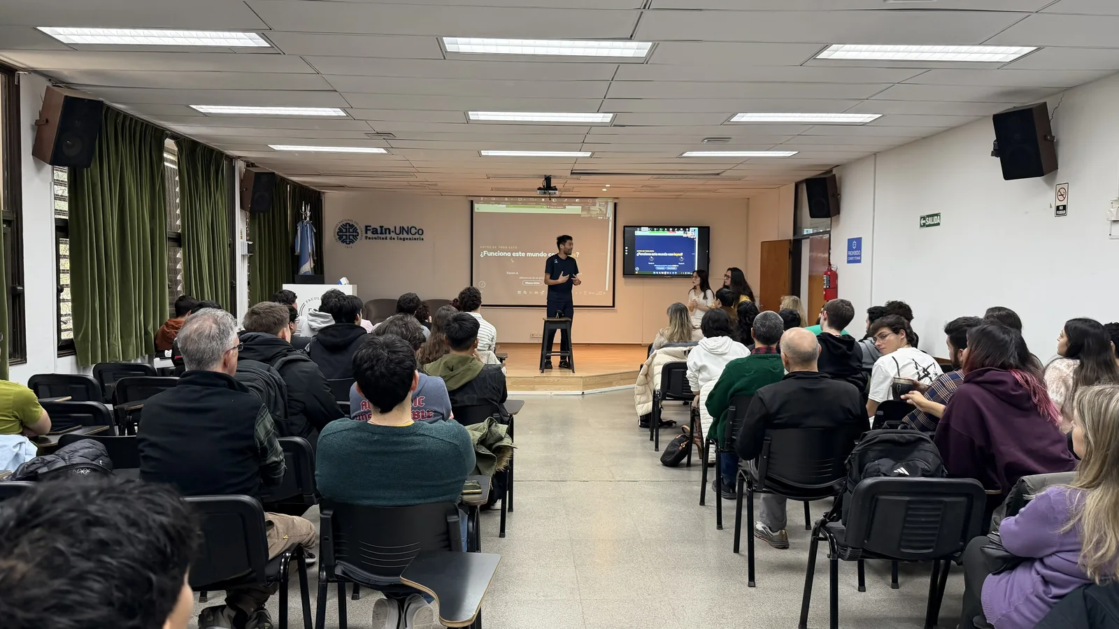 コマウエ国立大学工学部の講堂。壁に「FaIn·UNCo Facultad de Ingeniería」のロゴがあり、席を埋めた40人ほどの聴衆の前で、田中香津生が舞台に立って話している。背面のスクリーンにはスペイン語のスライド、その右のモニターにはオンライン参加者の画面が映っている
