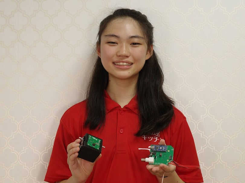 Yuzuka Sasaki smiling in a red Accel Kitchen polo shirt, holding a small cosmic-ray detector in each hand