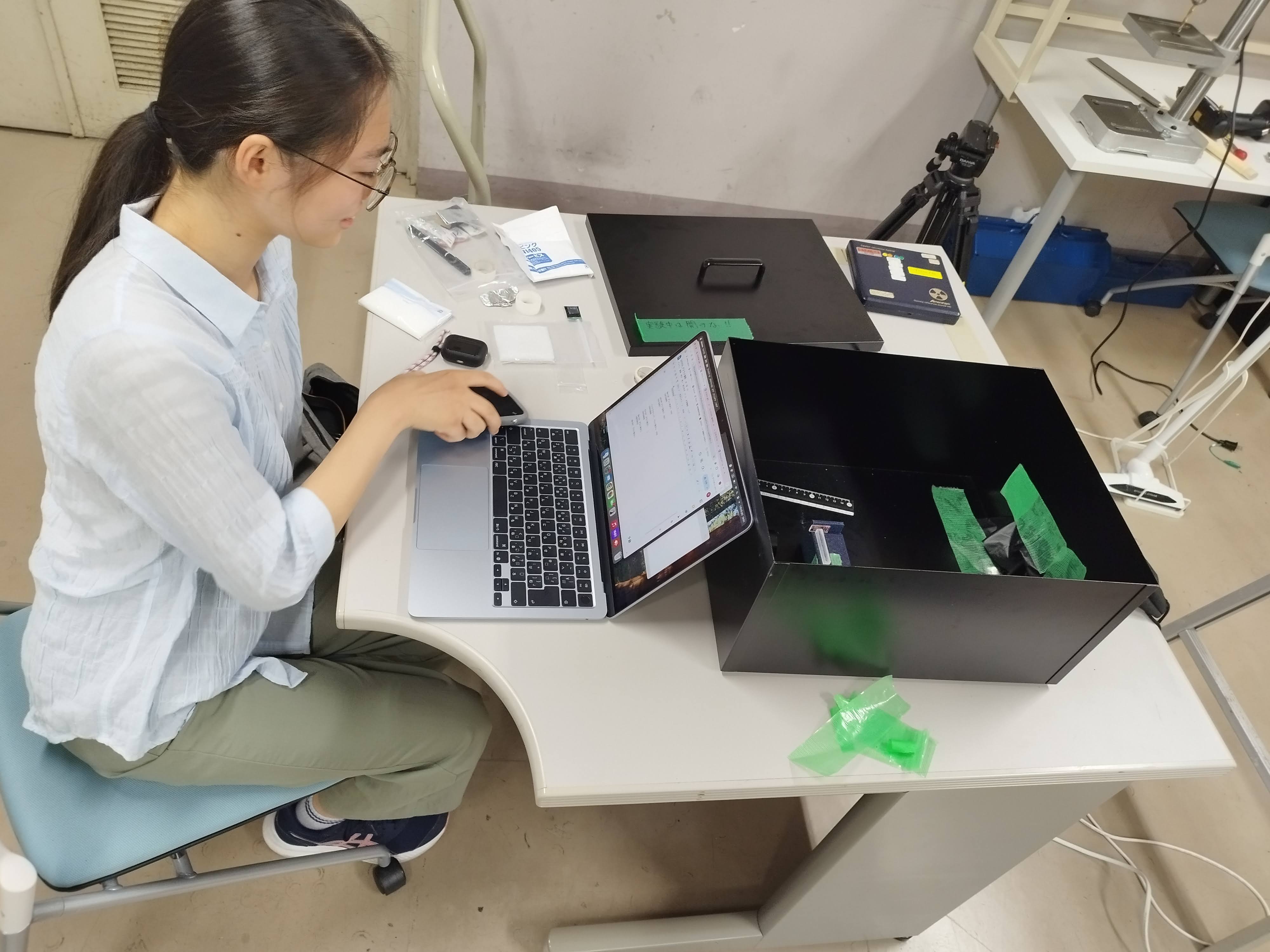 Yuzuka Sasaki at a lab bench with a laptop in front of her and, to the right, an opened black light-tight box containing a ruler and a scintillator standing on a blue mount; the lid carries a note reading 'Do not open during the experiment!!'