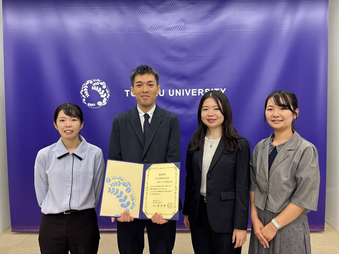 東北大学の紫色のバックパネルの前で、表彰状を手に並ぶ4人。開かれた表彰状には「第13回澤柳記念DEI賞 加速キッチン合同会社 殿」と記されている
