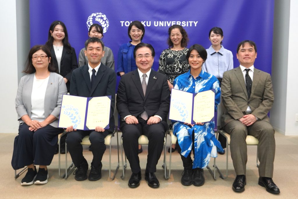東北大学の紫色のバックパネルの前で撮影した集合写真。前列に椅子に座った5人、後列に立った5人が並び、前列の2人が開いた表彰状を手にしている
