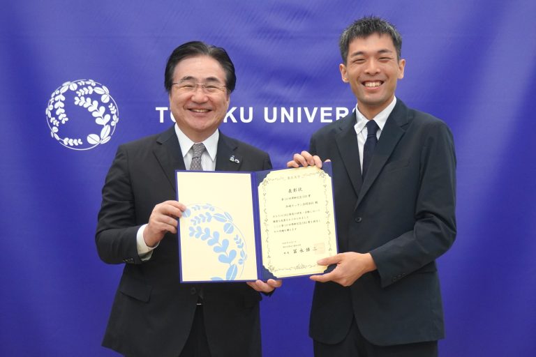 2人の男性が、東北大学の紫色のバックパネルの前で、開いた表彰状を両側から持ち、笑顔でカメラに向かっている